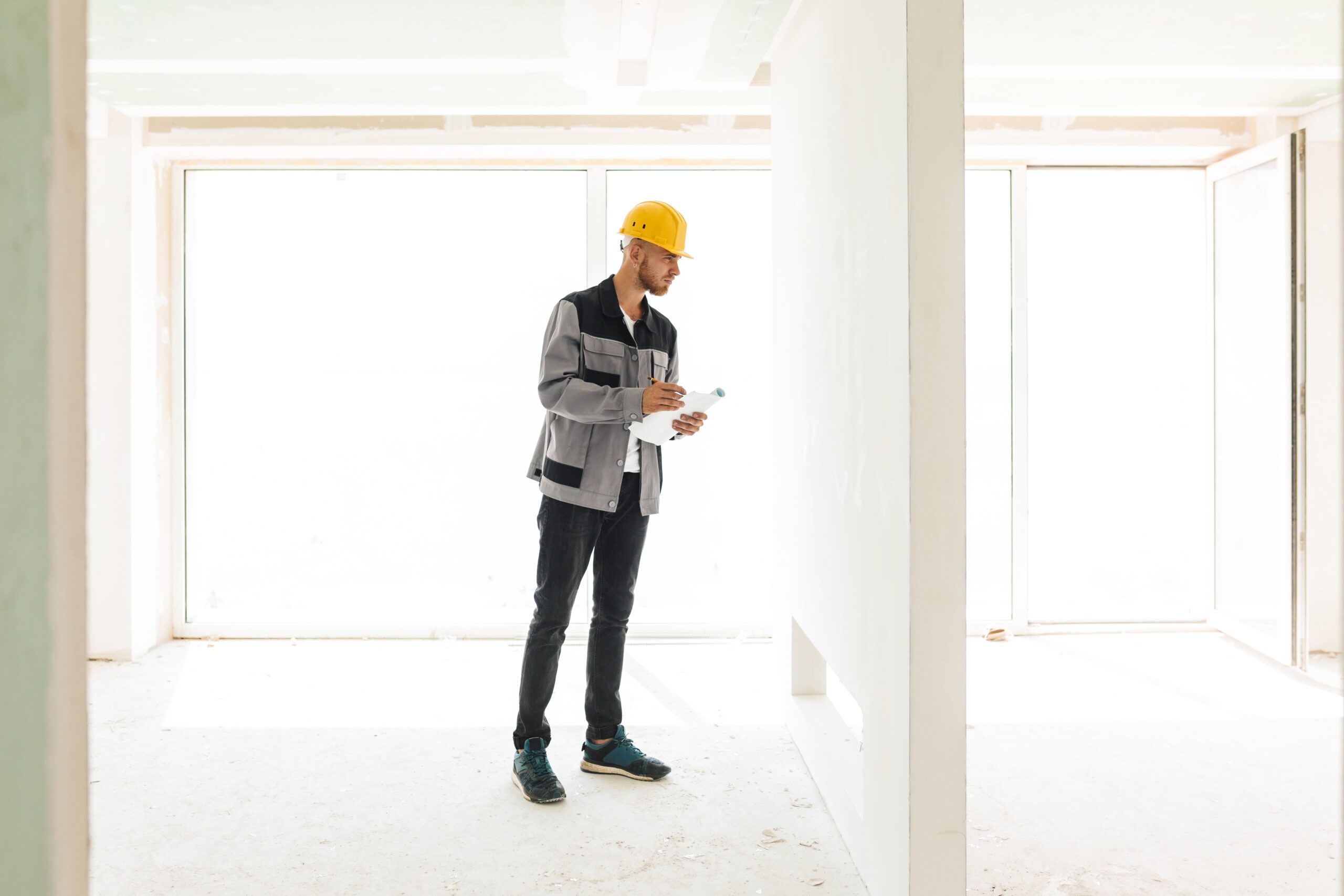 young-engineer-work-clothes-yellow-hardhat-holding-plan-new-apartments-thoughtfully-looking-wall-with-big-window-background-min